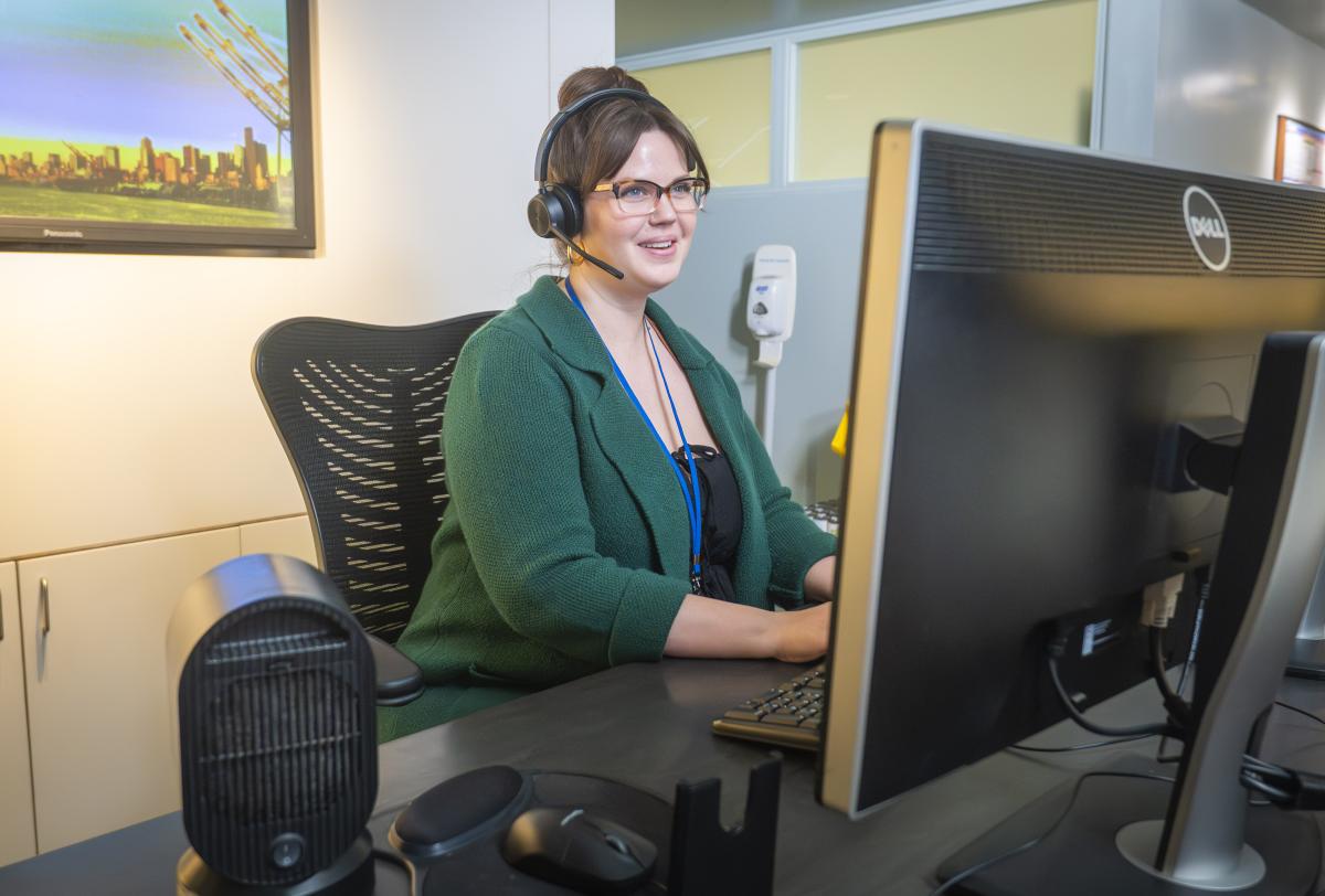 A woman in a green shirt wearing a headset types on a keyboard in front of a monitor.
